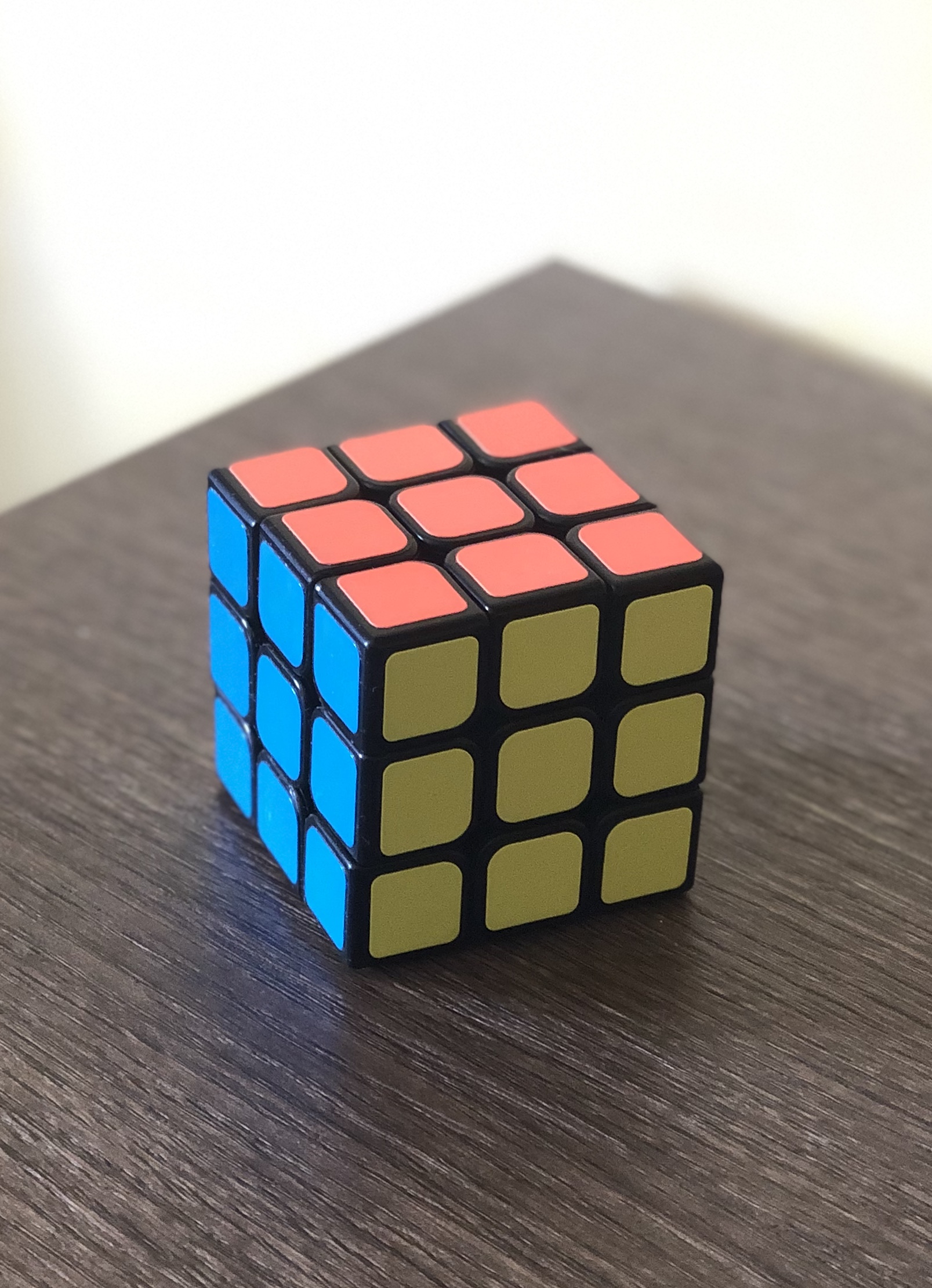 A solved three by three Rubik's Cube standing on a dark wooden table, one face entirely orange, the adjoining faces entirely olive and entirely blue