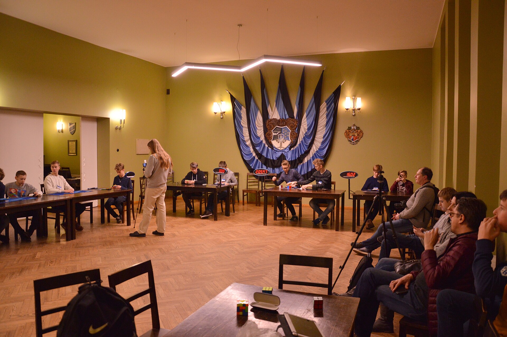 A competition hall with wood floors and olive walls, competitors seated at long tables in pairs with red digital timers in front of them, a judge standing in the middle of the room and an audience watching from chairs along one side