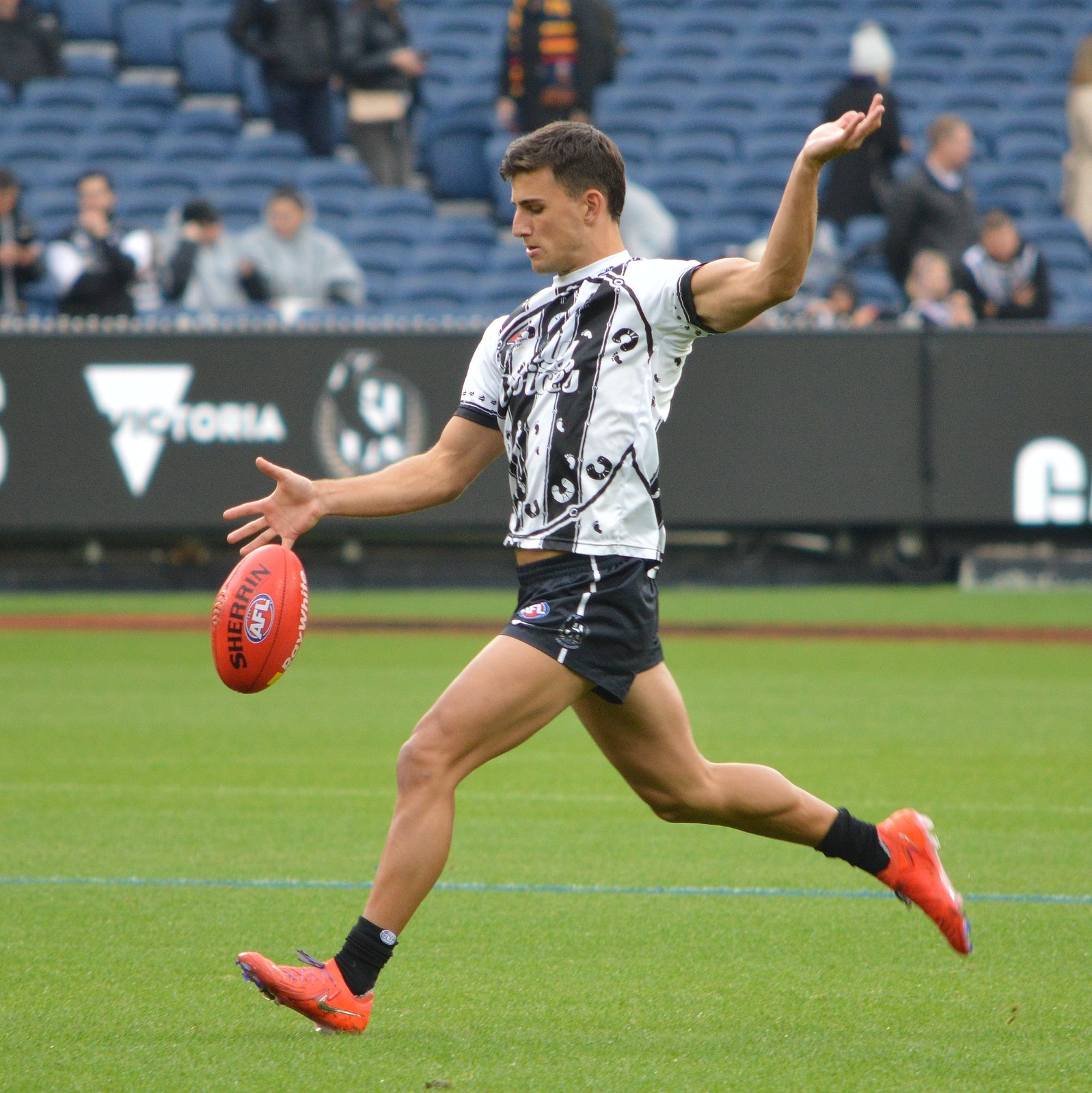 Nick Daicos in Collingwood's black and white clash guernsey, mid-stride and about to kick, dropping a red Sherrin towards his boot at the MCG
