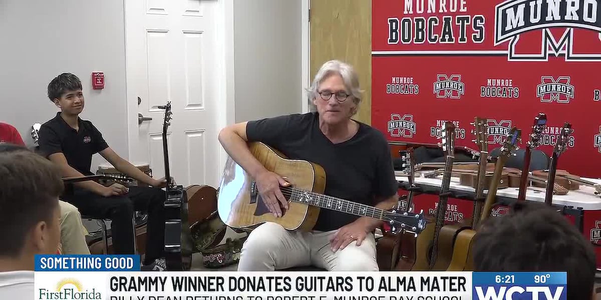 Billy Dean, grey-haired and in a black T-shirt, sits playing an acoustic guitar in front of a red Munroe Bobcats banner, with a row of new guitars on stands behind him and a boy holding a guitar and smiling at the left of the frame