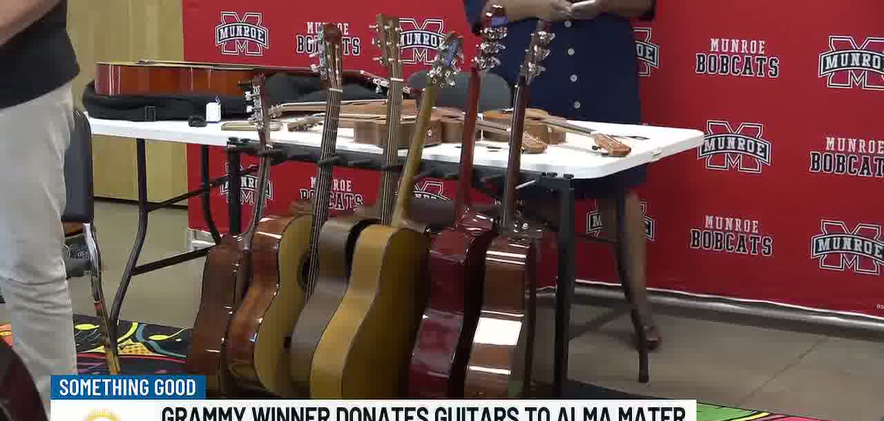 Ten new acoustic guitars on stands on a folding table in front of the red Munroe Bobcats banner, with a violin case on the table and a music-themed rug on the floor