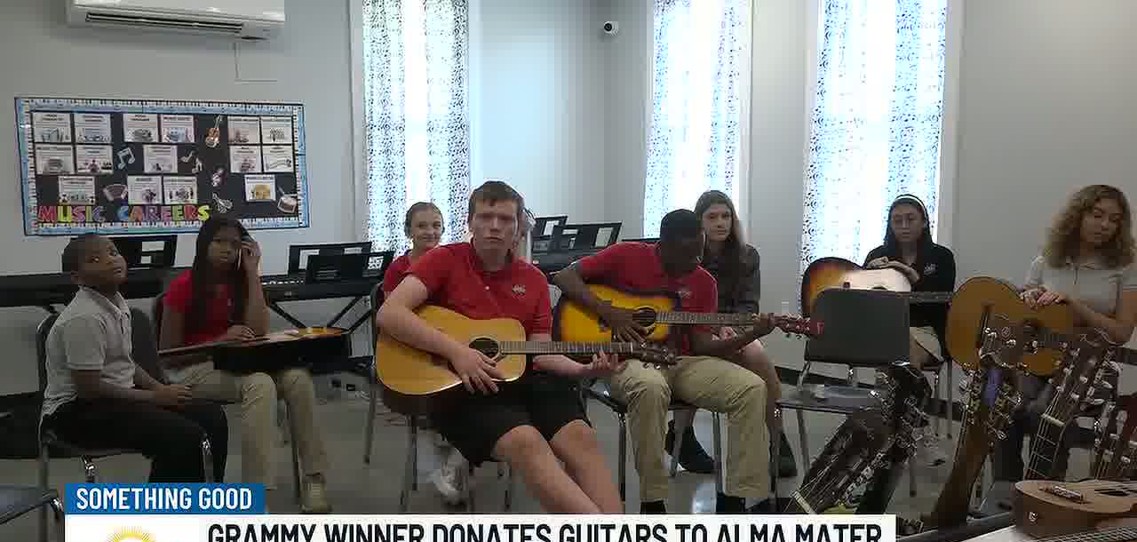 Students in red and white polo shirts sit in a classroom holding acoustic guitars, one boy in the middle playing, with more guitars on stands in the foreground and a Music Careers display on the wall