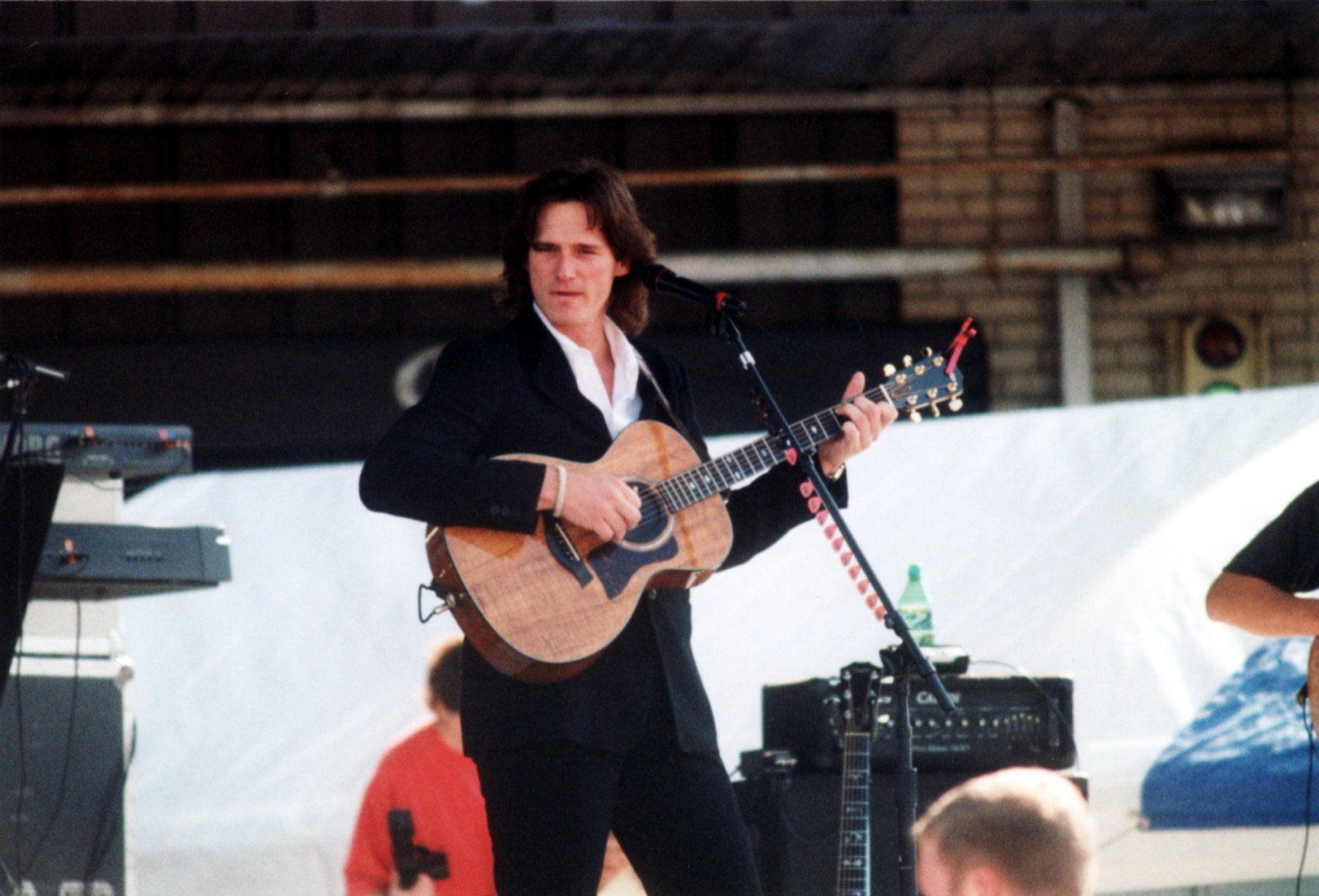 Billy Dean in 1998, dark-haired and in a black suit with an open white shirt, playing an acoustic guitar at a microphone on an outdoor stage
