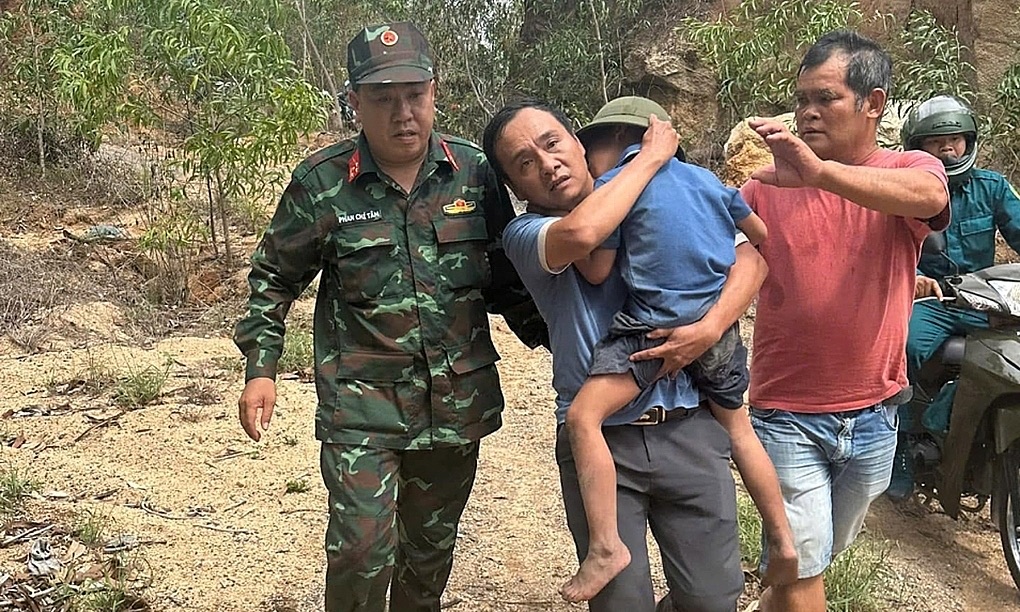 A man in a blue shirt carries a barefoot six-year-old boy down a sandy forest track, a soldier in camouflage walking beside him and another man in a red shirt behind, with a militia member on a motorbike following