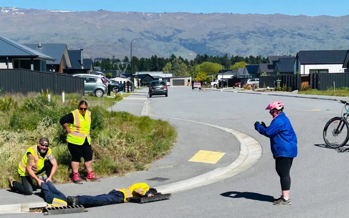 Two road workers in hi-vis vests by a drain at the kerb, one lying flat on the road reaching into it, while a cyclist in a blue jacket watches.
