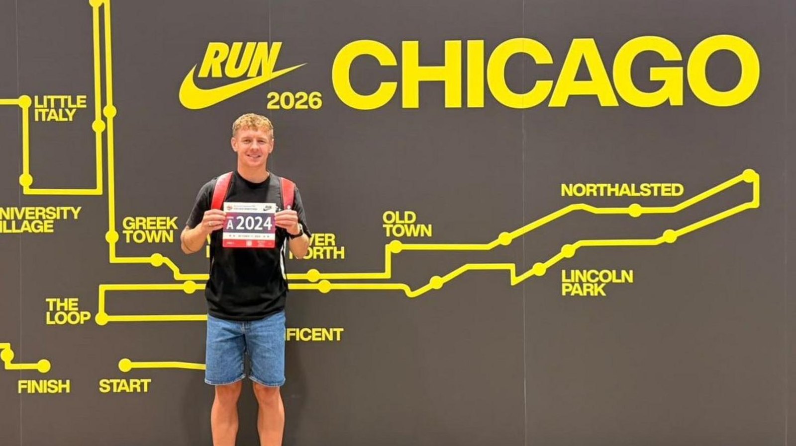 A young man with curly fair hair in a black T-shirt, denim shorts and a red rucksack, smiling and holding up a race number, A2024, in front of a wall printed with the Chicago Marathon route map.