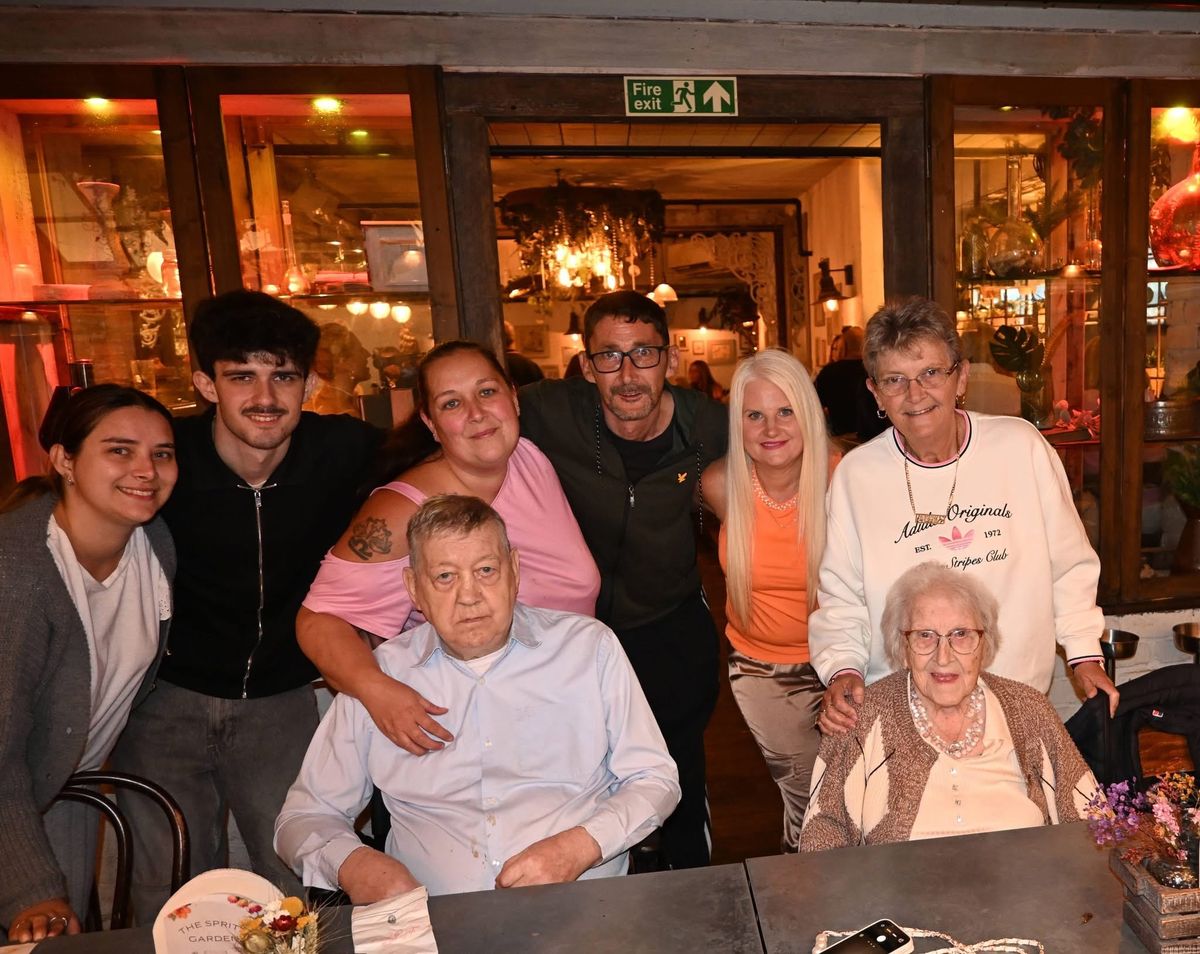 Seven members of a family standing behind William and Susan, who are seated at a restaurant table, all facing the camera.