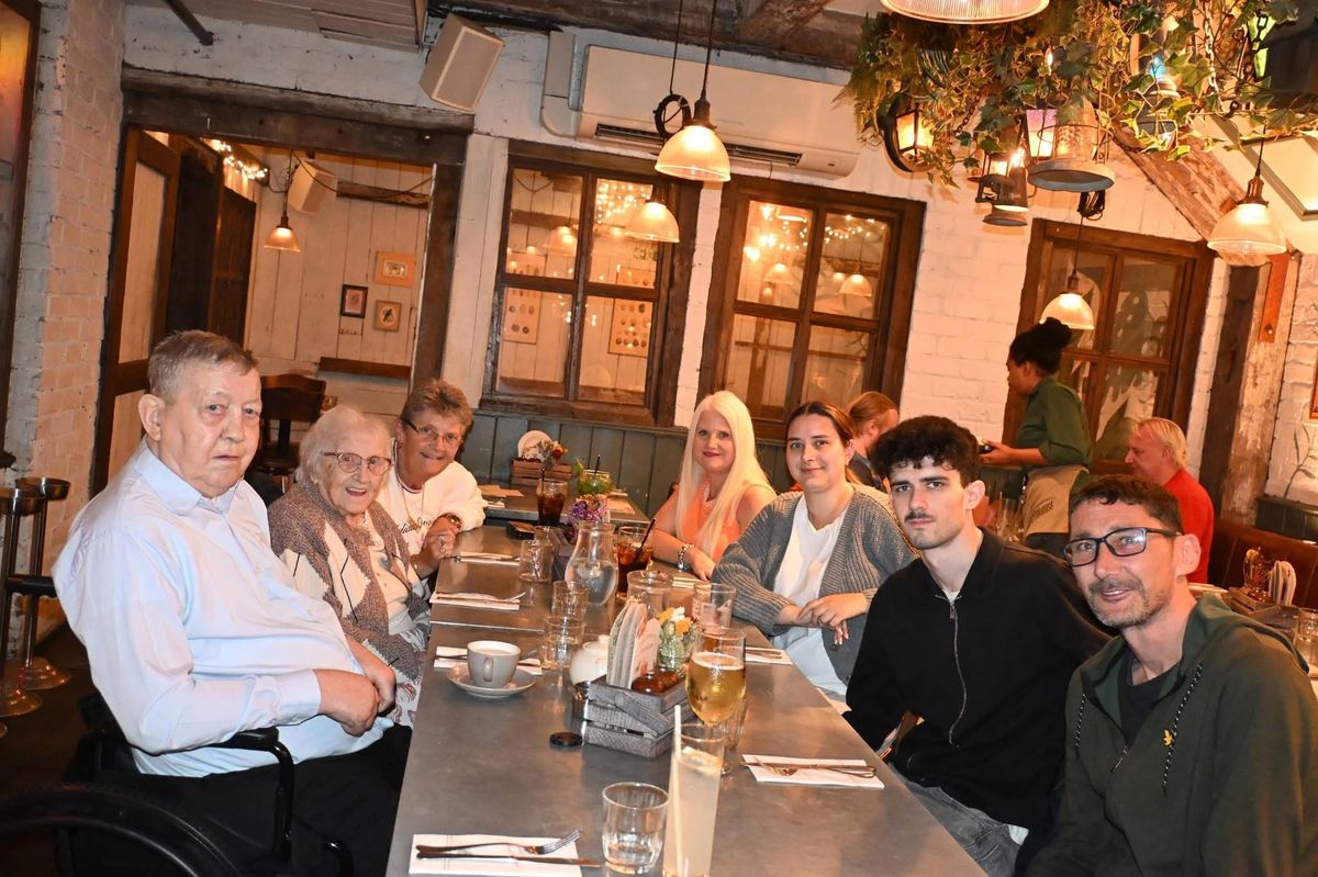 A long restaurant table under hanging lamps with William, Susan and their family seated along both sides, looking towards the camera.