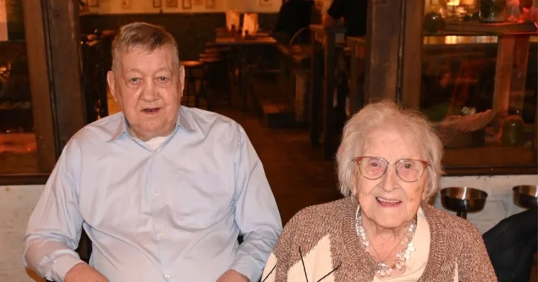 An elderly man in a pale blue shirt and an elderly woman in glasses, a beaded necklace and a knitted cardigan, sitting side by side at a restaurant table and smiling.