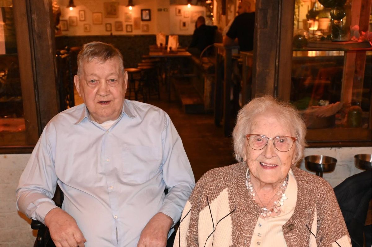 An elderly man in a pale blue shirt and an elderly woman in glasses, a beaded necklace and a knitted cardigan, sitting side by side at a restaurant table and smiling.