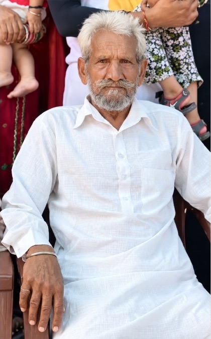 Chaman Lal seated in a chair in a white kurta, white hair combed back and a white beard, a silver bangle on his wrist, family members standing behind him