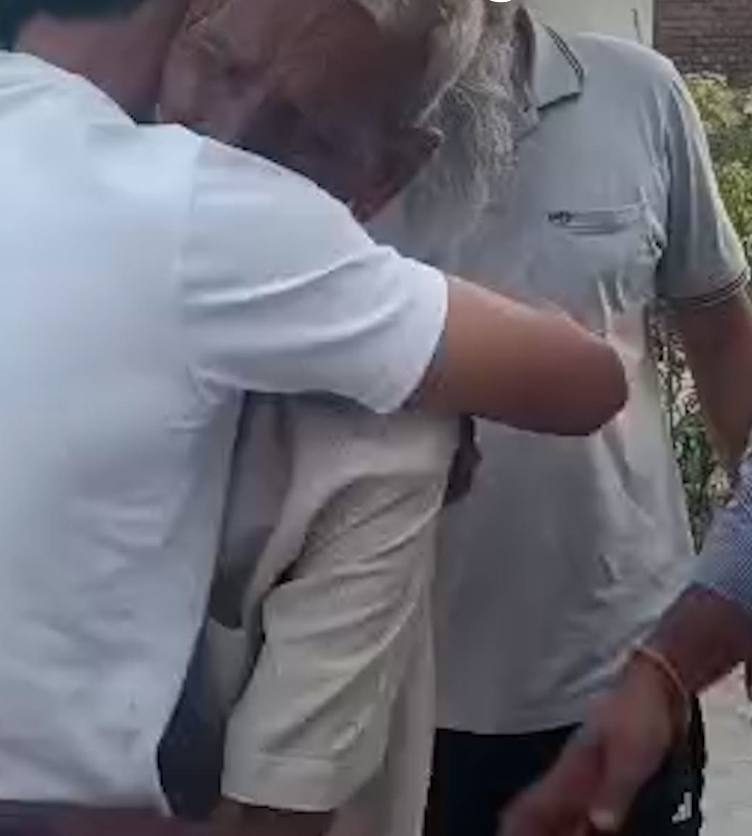 A young man in a white t-shirt holds an elderly man with a long white beard in a tight embrace in a village street, other men standing close around them