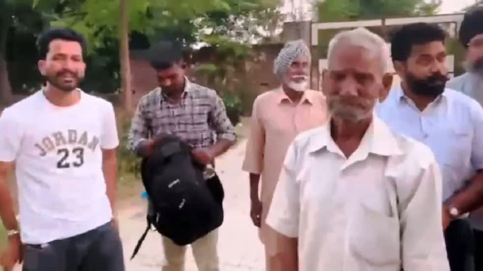 An elderly man with white hair and a white beard in a white kurta stands in a village lane in front of a group of men, one of them a young man in a checked shirt holding a black backpack, another in a white Jordan t-shirt, a man in a turban behind them