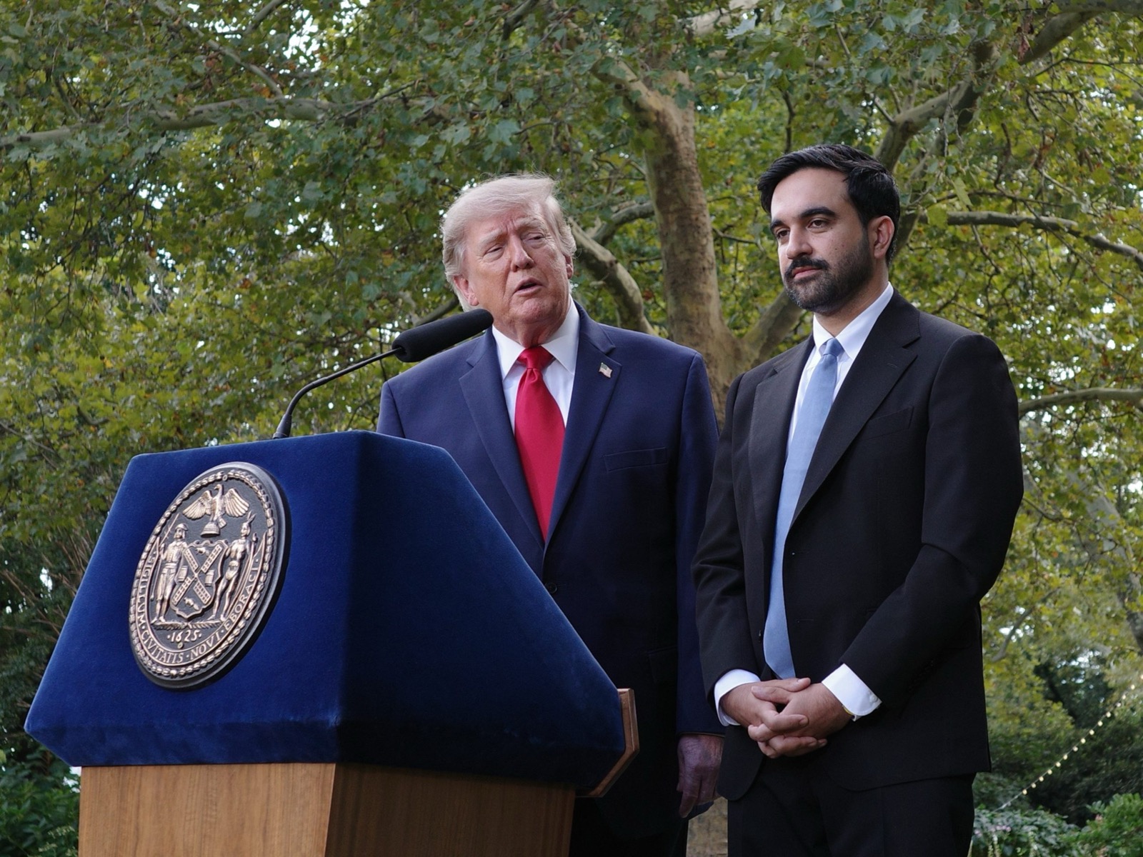 Donald Trump speaks at a blue lectern carrying the seal of the City of New York on the lawn of Gracie Mansion, with Zohran Mamdani standing beside him in a dark suit and pale blue tie, his hands clasped, under the trees