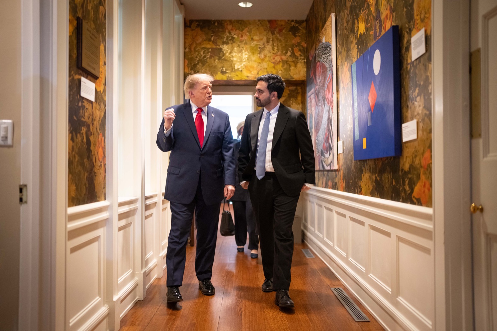 Trump and Mamdani walk side by side down a narrow hallway inside Gracie Mansion, past patterned wallpaper and framed paintings, Trump in a navy suit and red tie gesturing with one hand, Mamdani in a dark suit listening