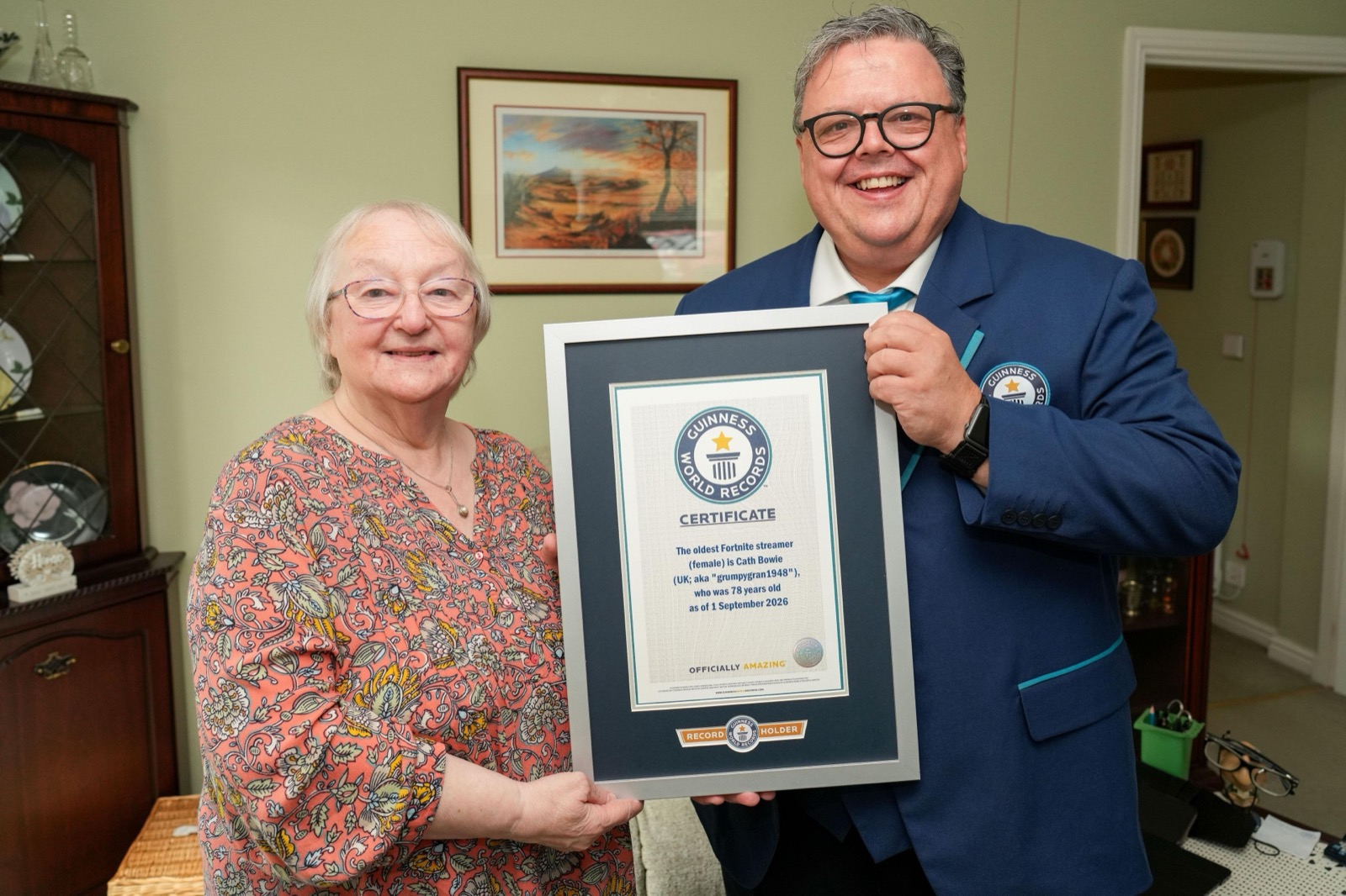 The woman stands in a living room holding a framed Guinness World Records certificate with an adjudicator in a blue blazer, both smiling at the camera