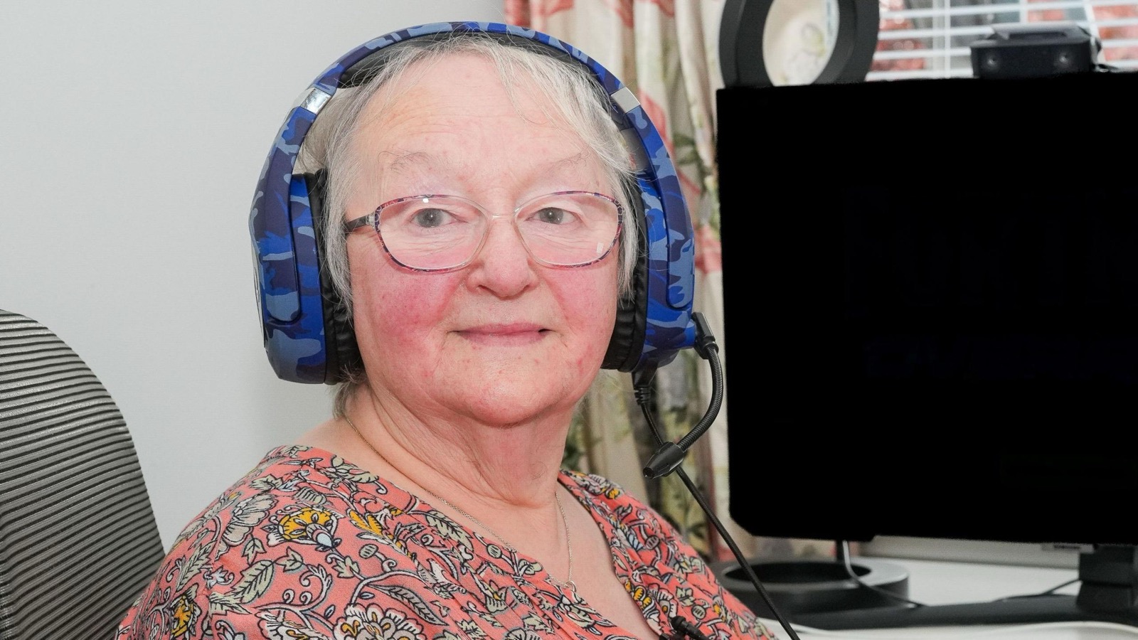A woman with short grey hair and glasses wearing a blue camouflage gaming headset with a boom microphone, sitting beside a monitor and a webcam, smiling at the camera