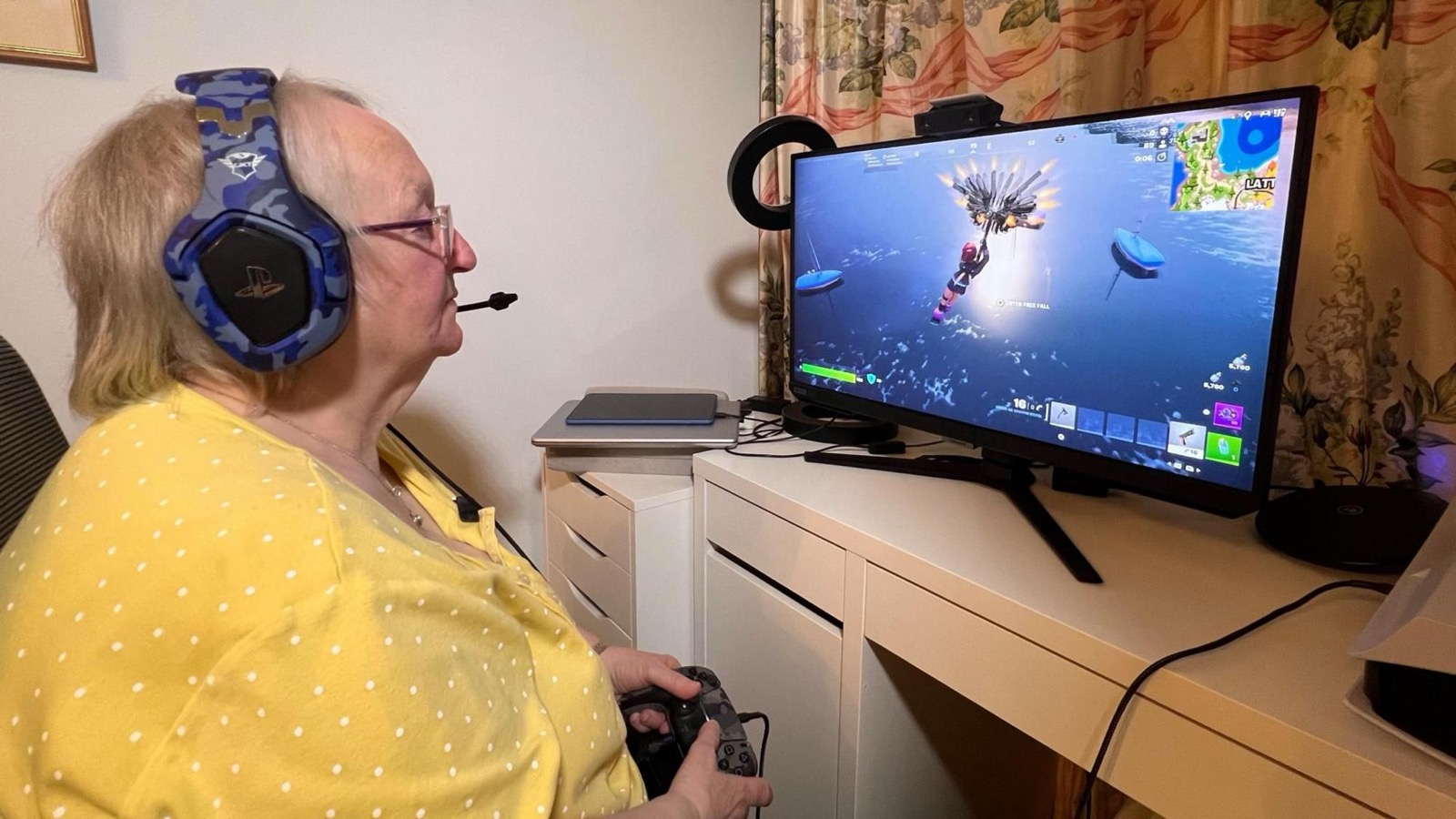 The same woman in a yellow polka-dot top and a PlayStation headset, controller in both hands, watching a monitor on which a Fortnite character parachutes towards water