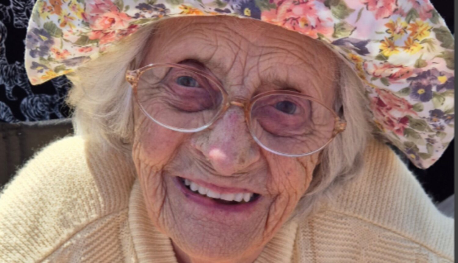 Gwen Chapman smiles in large glasses, a floral sun hat and a cream cardigan