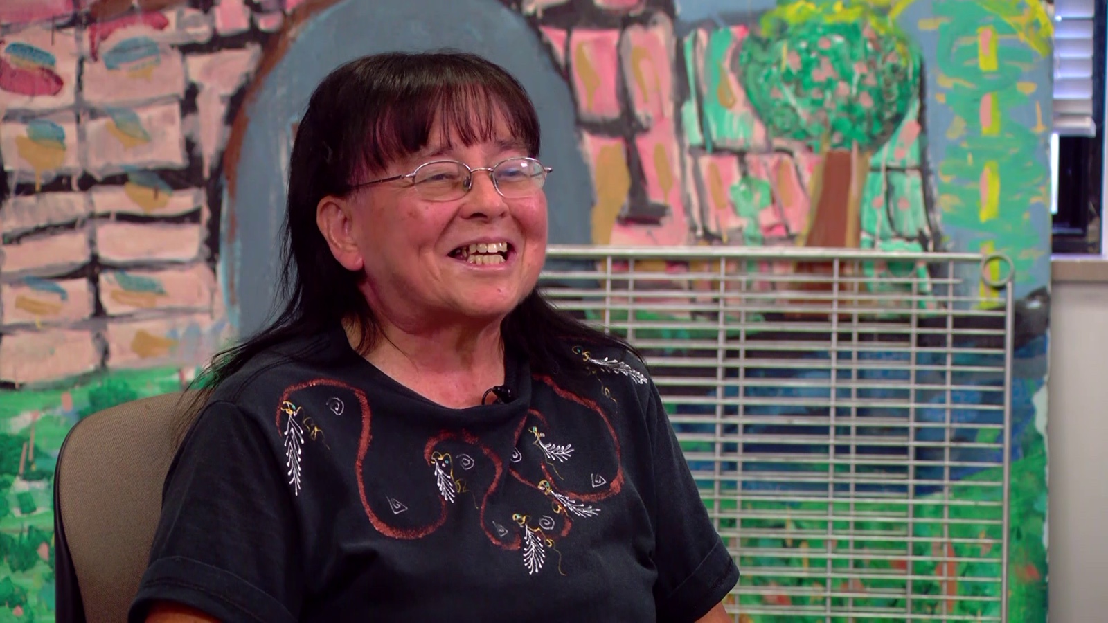 Marcelina Agbuis, in glasses and a dark embroidered top, smiling, sitting in front of the same painted mural