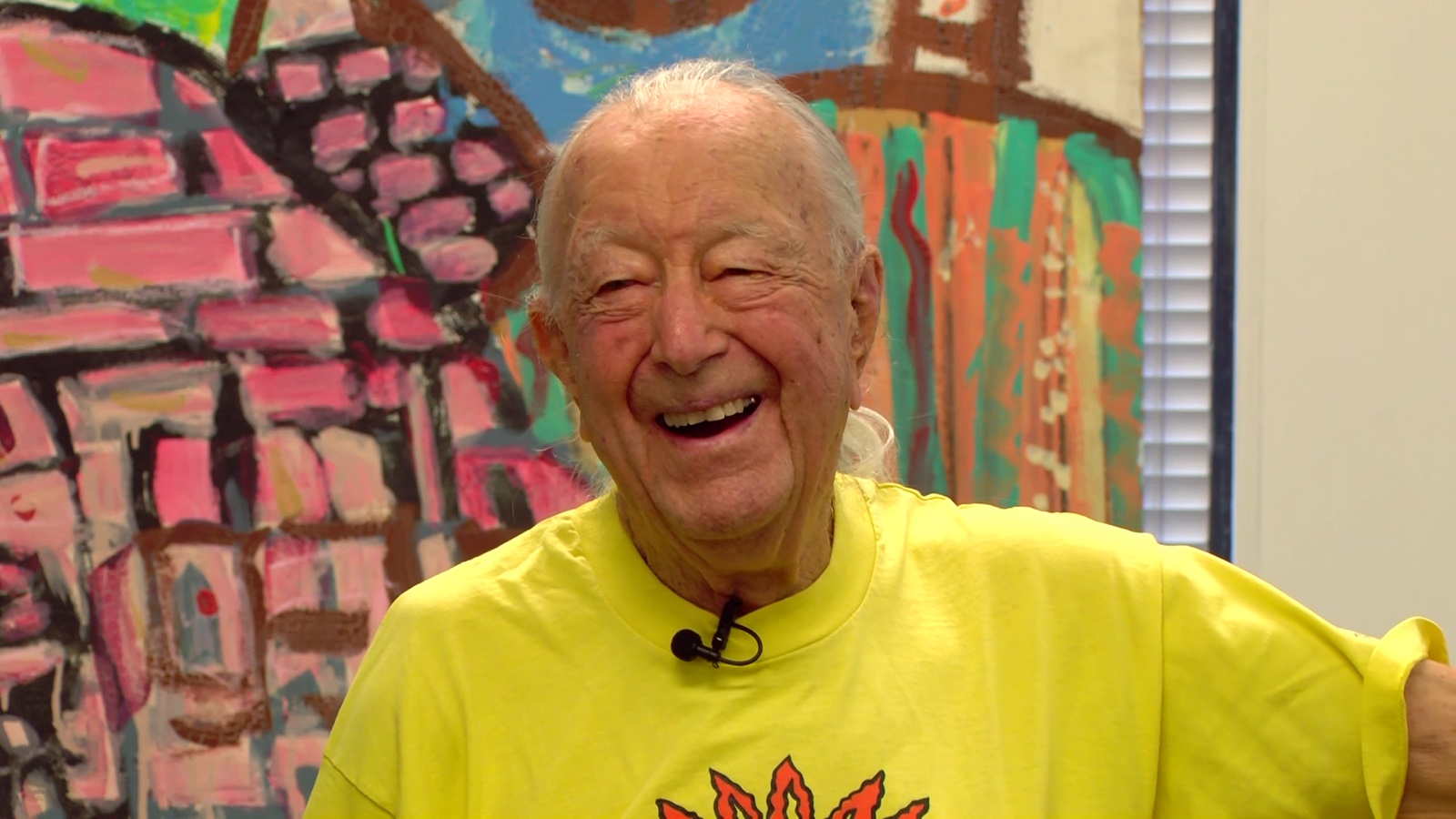 Dick Knepper, laughing, in a yellow T-shirt with a clip-on microphone, sitting in front of a brightly painted mural