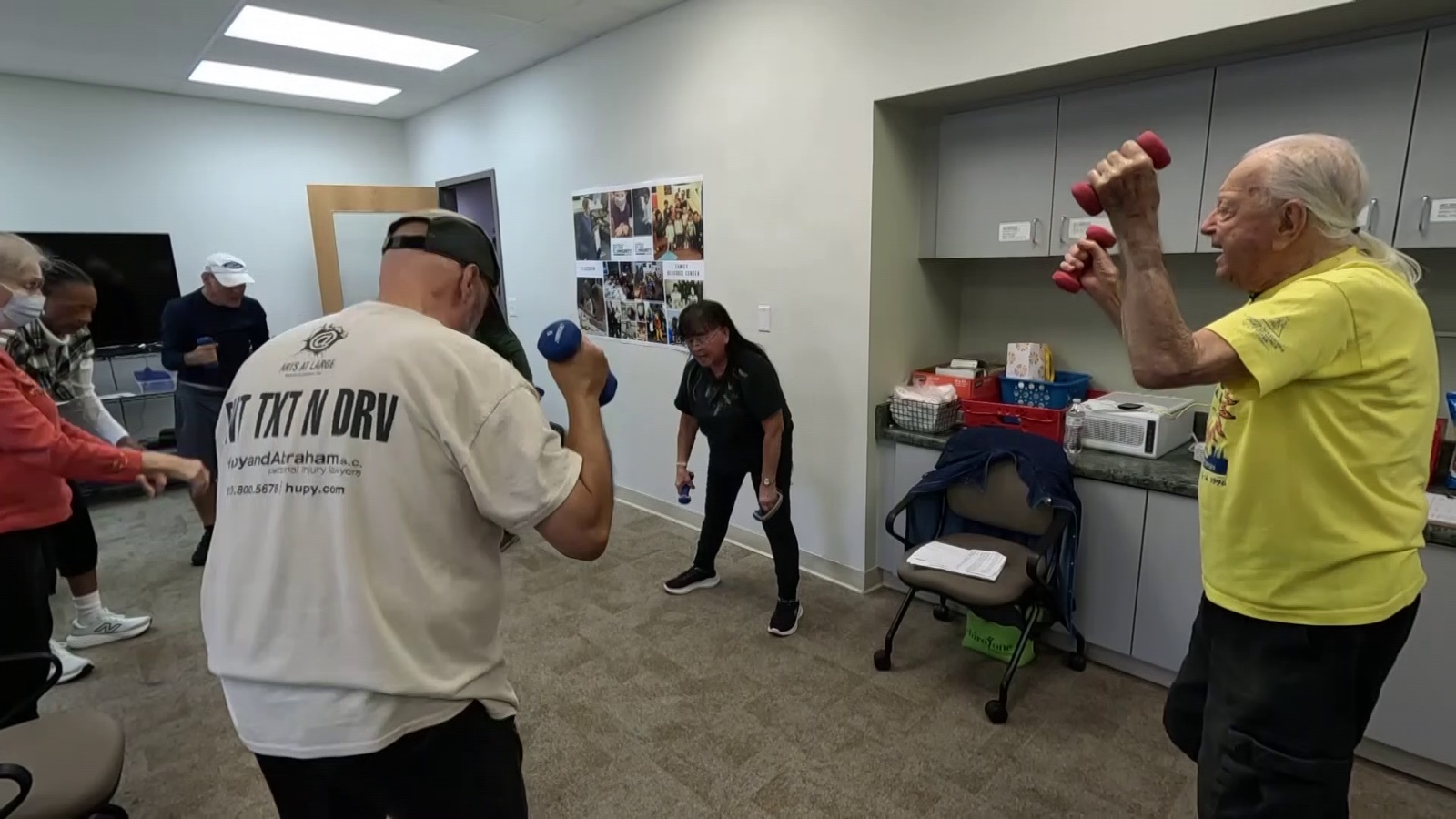 Dick Knepper in a yellow T-shirt pressing two red dumbbells above his head, facing a room where other people are lifting smaller weights