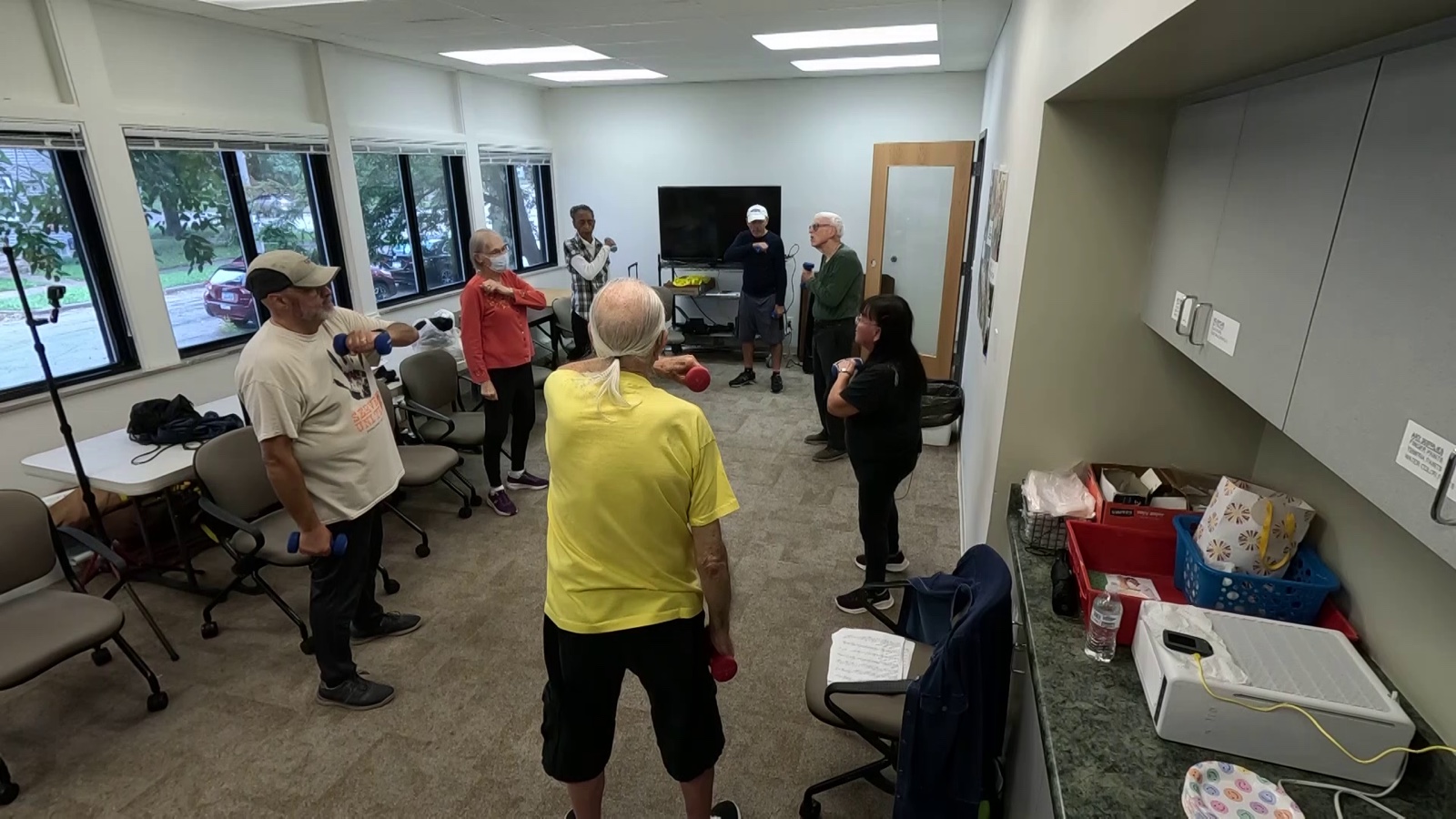 A community centre room with windows along one wall, about eight people standing in a loose circle with dumbbells, Knepper in his yellow shirt leading them