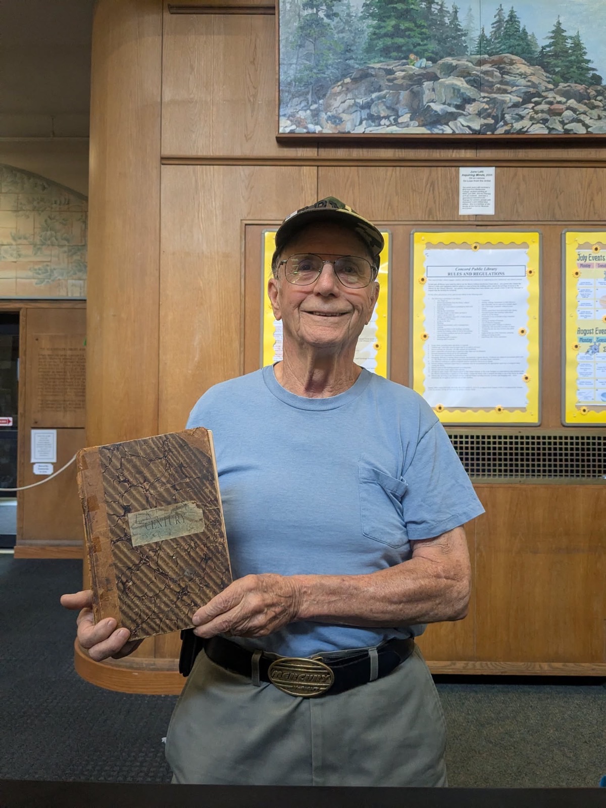 John Hanson, in a camouflage cap, glasses and a pale blue t-shirt, holds a worn marbled hardback with a label reading Century, standing in the Concord Public Library lobby in front of a noticeboard headed Rules and Regulations