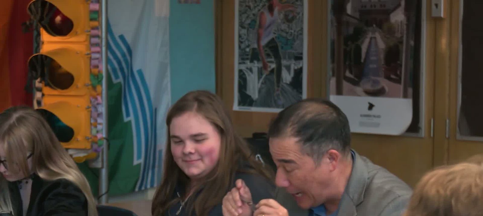 Steve Hobbs leaning in at a desk beside a smiling student, both laughing, a traffic light and posters behind them.