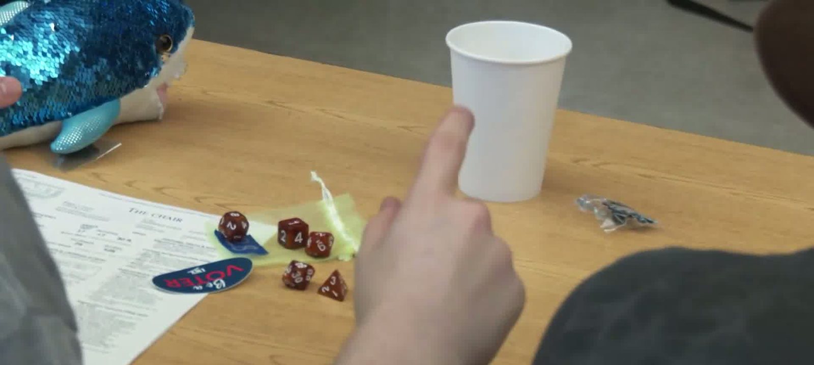 Red and gold polyhedral dice and a character sheet on a desk beside a paper cup and a round sticker that reads Vote.