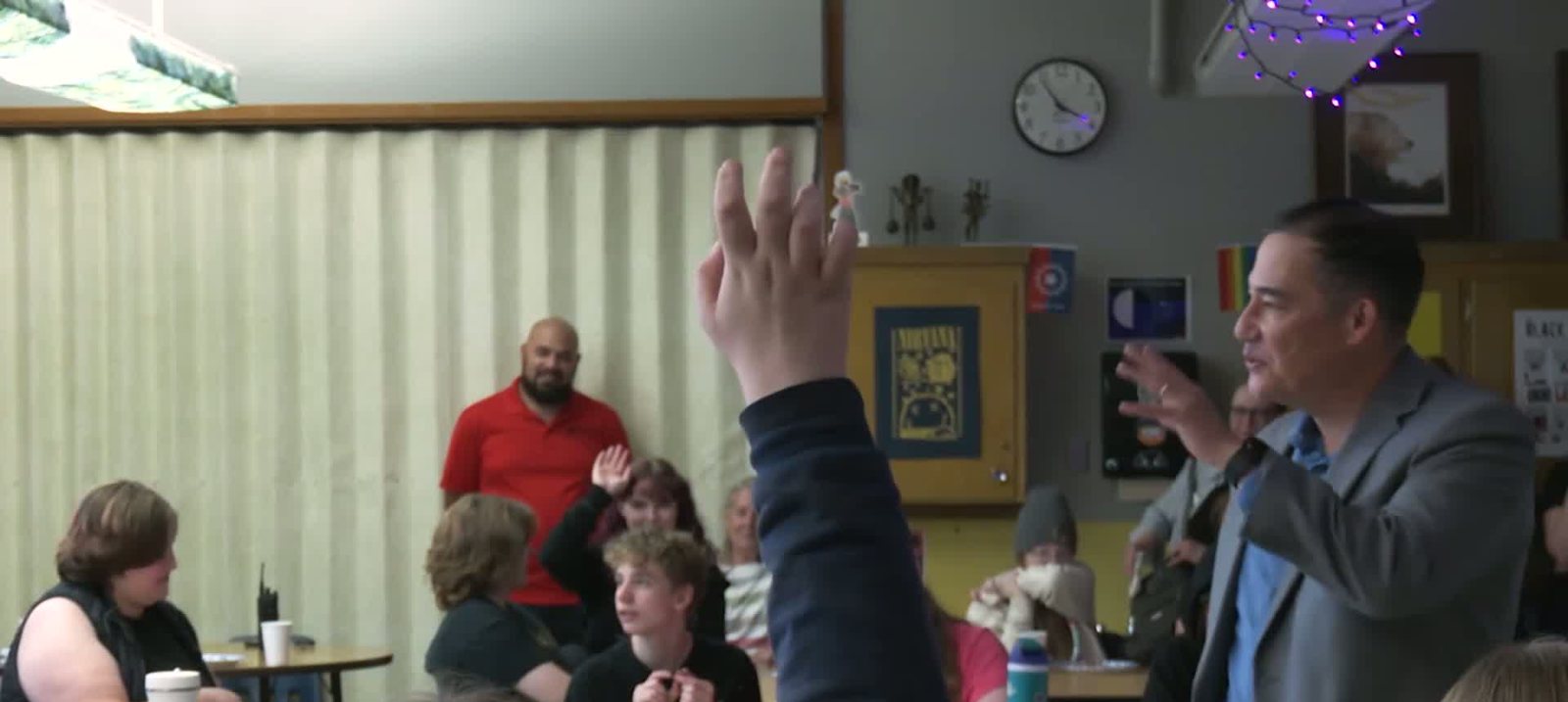 A student&rsquo;s raised hand in the foreground while Steve Hobbs, in a grey jacket, gestures as he answers questions in a classroom with string lights and posters.