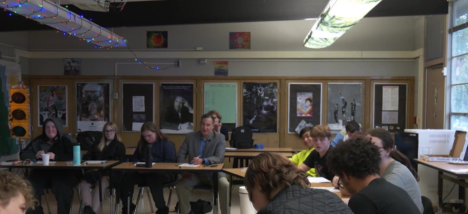 Steve Hobbs in a grey jacket sitting at a row of classroom desks with high school students, dice and character sheets on the tables, string lights on the ceiling and posters on the wall behind.