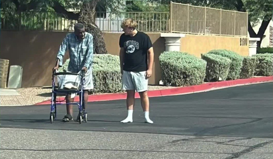 A teenager in a black T-shirt and white socks, no shoes, standing in the road beside an older man in a checked shirt who is wearing slides and leaning on a blue walker.