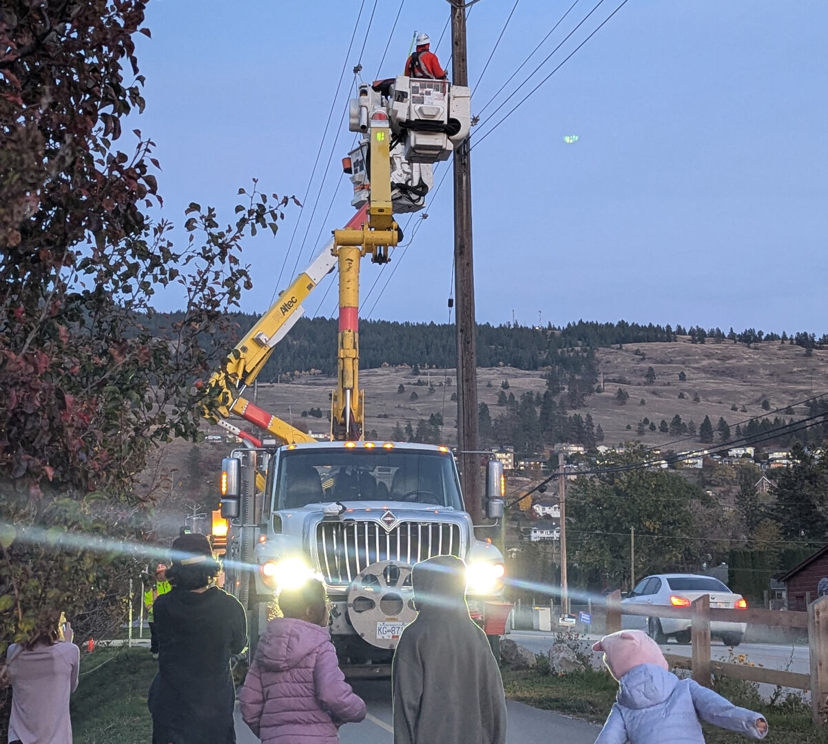 At dusk, a BC Hydro bucket truck with its lights on raises a worker up beside a power pole, while a few people watch from the roadside.