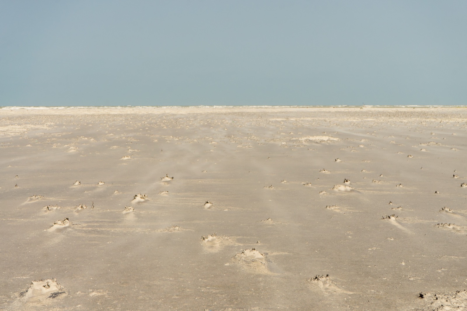 A wide empty beach of pale sand at Talaimannar in Sri Lanka, with low hummocks across it and the sea breaking in a thin white line along the far edge under a clear sky