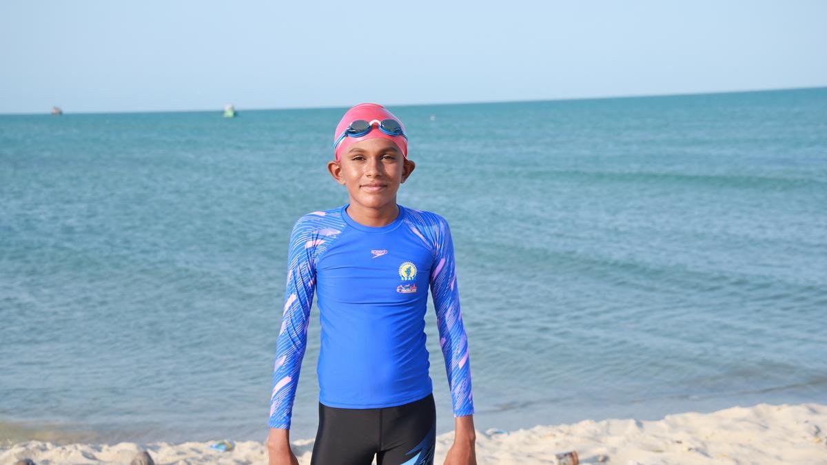 Ishaan, 13, standing on a white sand beach in a blue and pink long-sleeved swimming top and black leggings, a pink swimming cap on his head with goggles pushed up on it, calm turquoise sea behind him