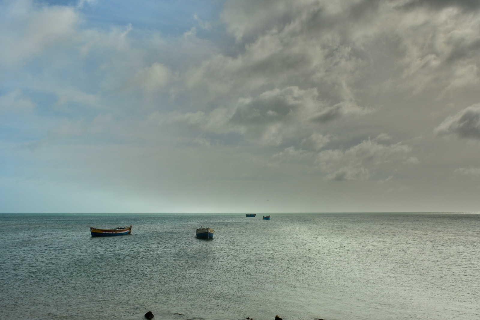 Flat grey-green sea at Dhanushkodi under a cloudy sky, with four small wooden fishing boats moored offshore and no land visible on the horizon