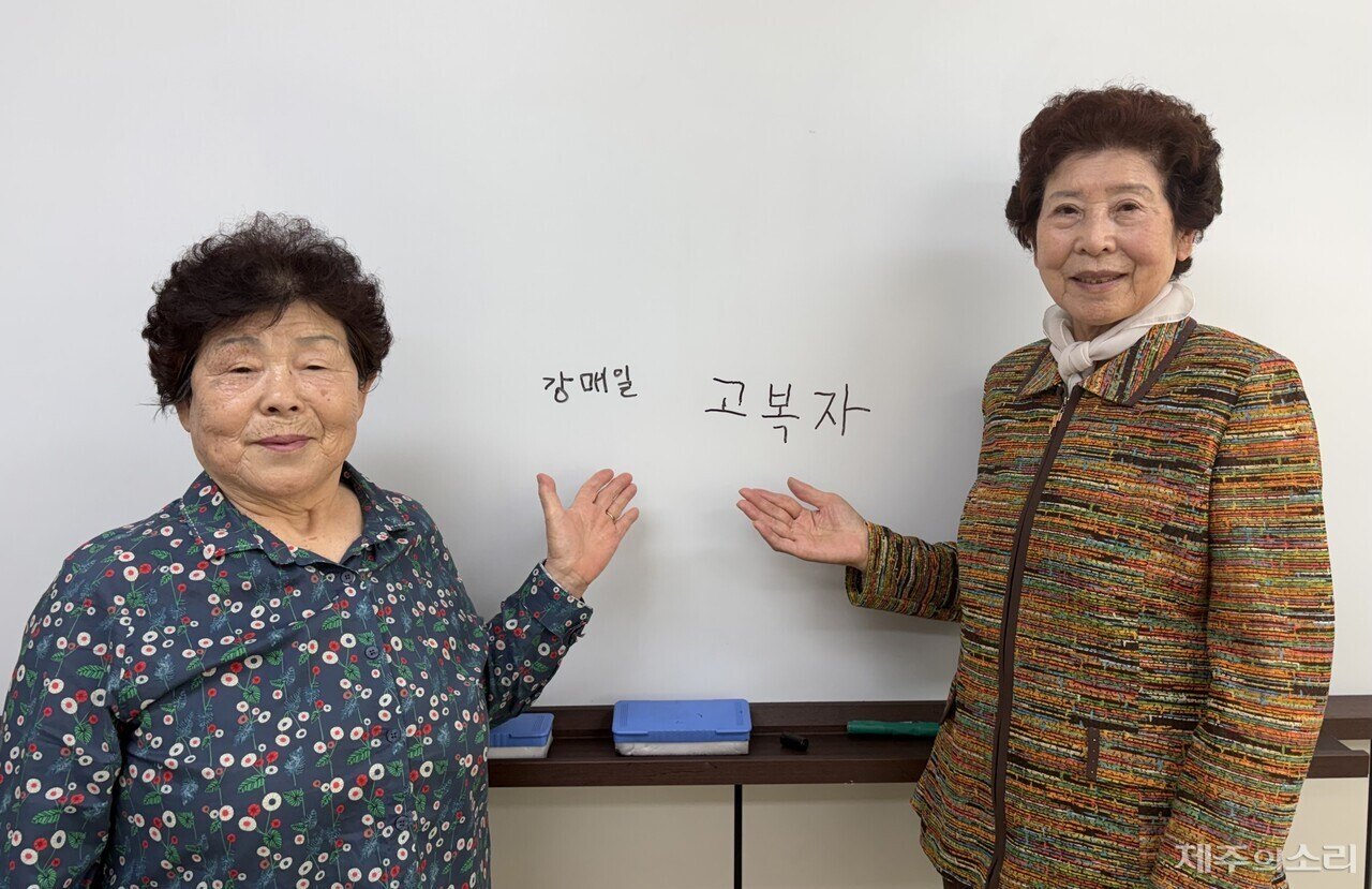 Two older Korean women in front of a whiteboard, each pointing at her own name written on it in Korean.