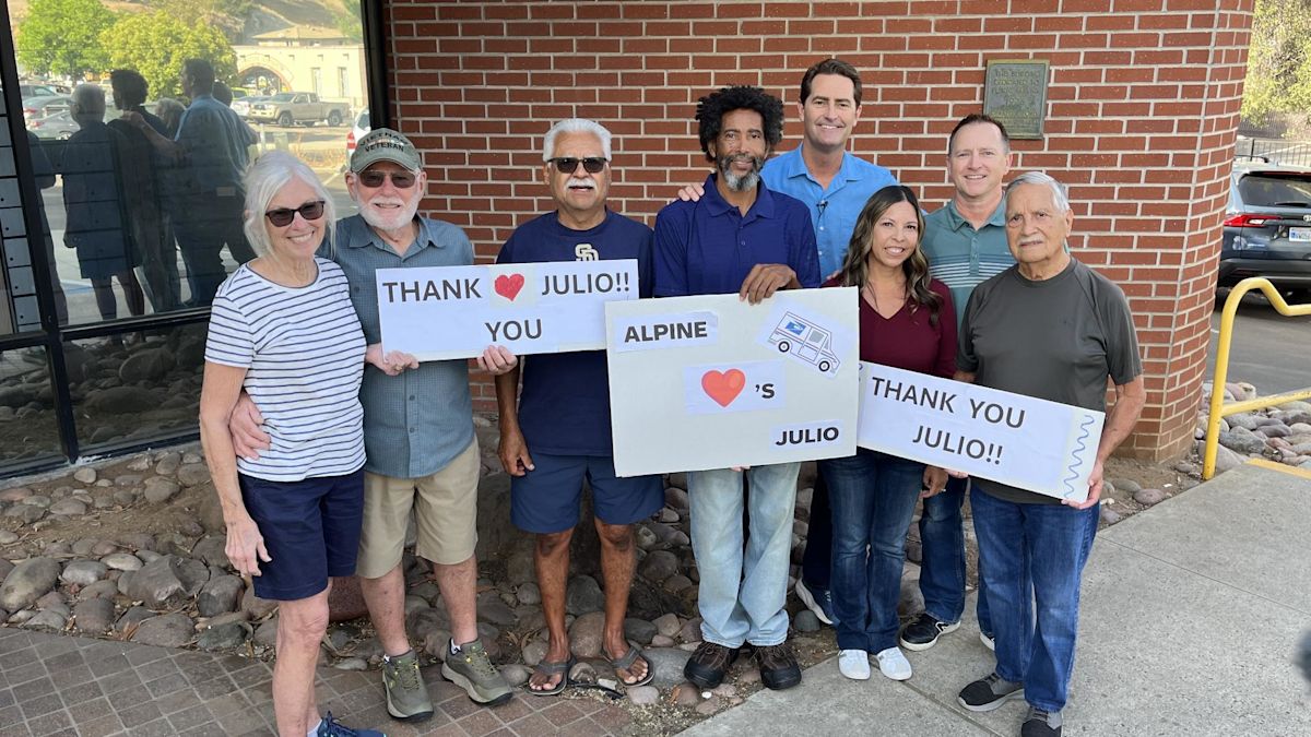 Eight people stand outside a brick post office holding hand-made signs that read Thank you Julio and Alpine loves Julio.