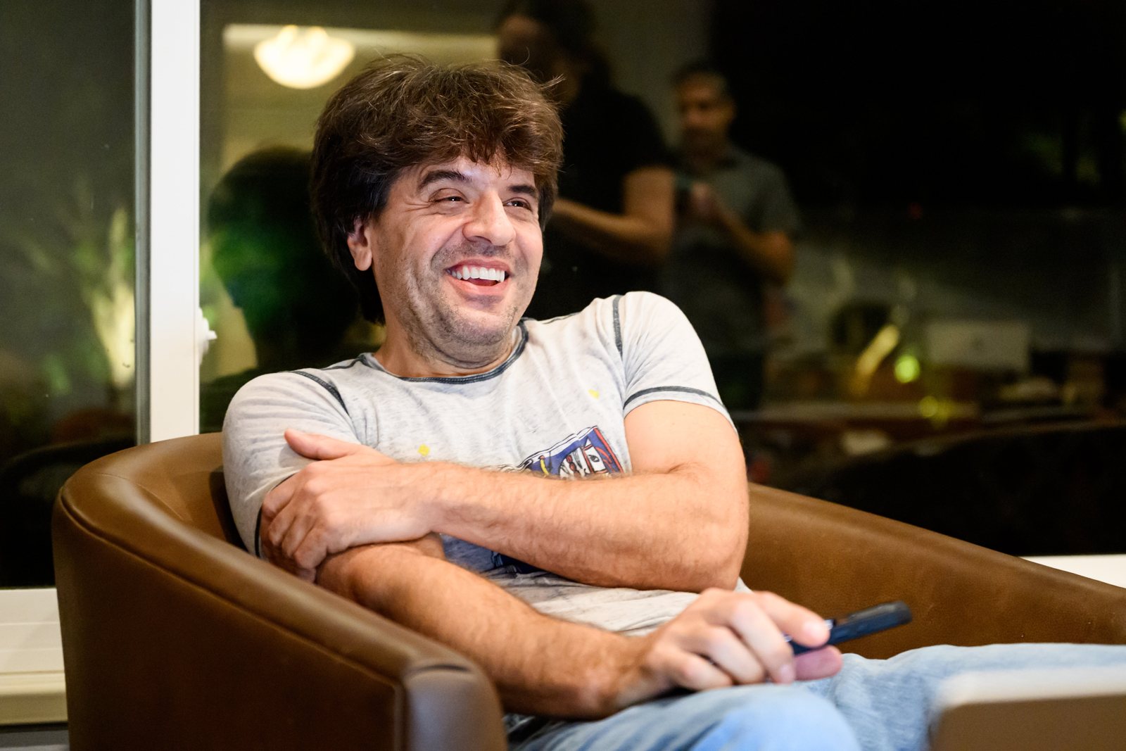 Karl Deisseroth laughing in a brown leather armchair at home at night, arms folded, wearing a grey T-shirt.