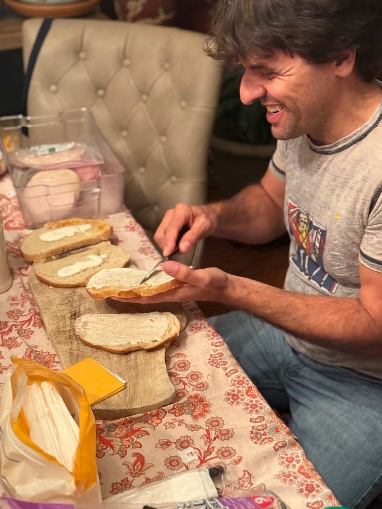 Karl Deisseroth, in a grey T-shirt, spreading filling on slices of bread laid out on a wooden board on a patterned tablecloth, with a slice of cheddar beside them.