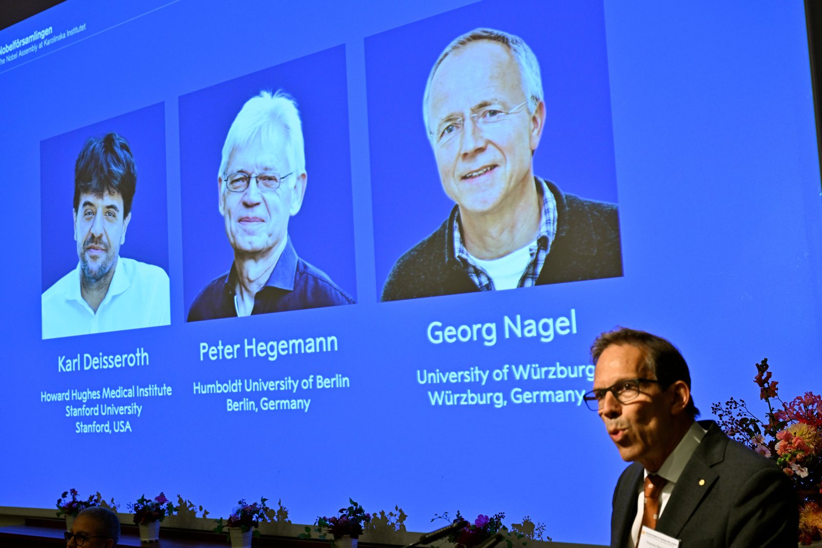 A large blue screen in Stockholm showing portraits of the three winners, Karl Deisseroth, Peter Hegemann and Georg Nagel, above a man speaking at a lectern.