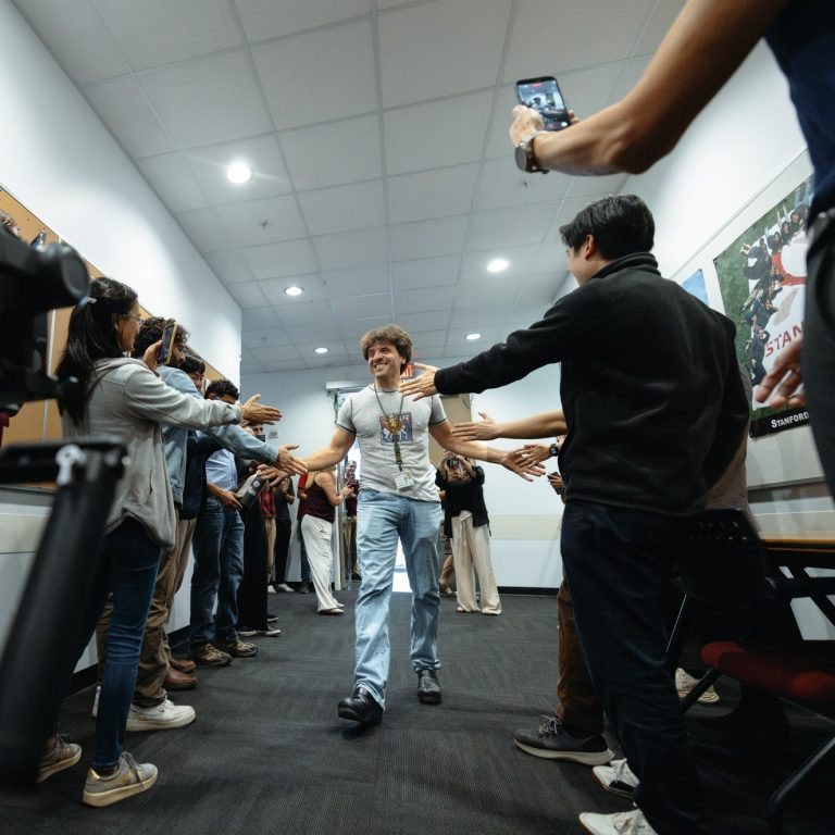 Karl Deisseroth walking down a corridor between two lines of colleagues holding out their hands to high-five him, with phones raised to film him.