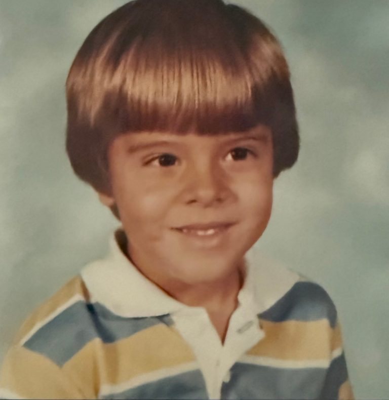 A school photograph of a young boy with a bowl haircut and a striped polo shirt, smiling.