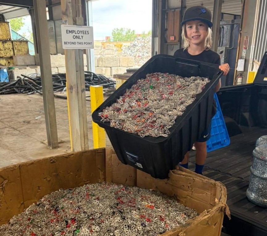 A smiling boy with long fair hair and a black cap tips a black tote full of pop tabs into a large cardboard bin of tabs at a recycling yard.