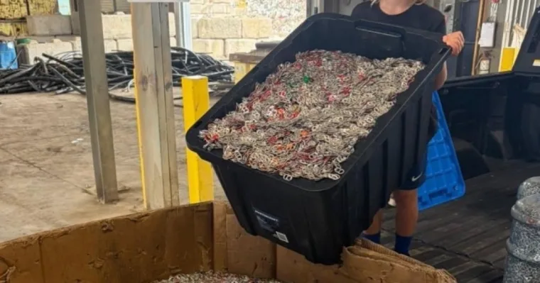 A smiling boy with long fair hair and a black cap tips a black tote full of pop tabs into a large cardboard bin of tabs at a recycling yard.