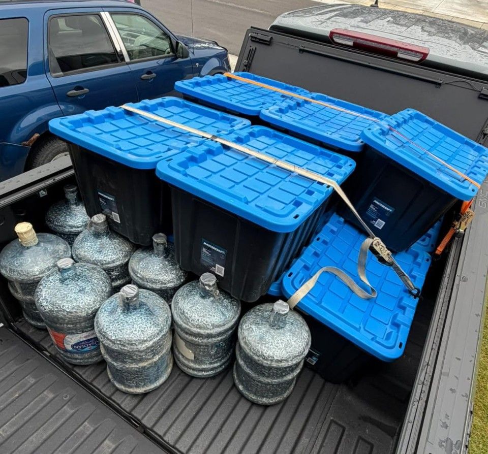 The bed of a pickup truck packed with black totes with blue lids and large water jugs filled with pop tabs, strapped down.