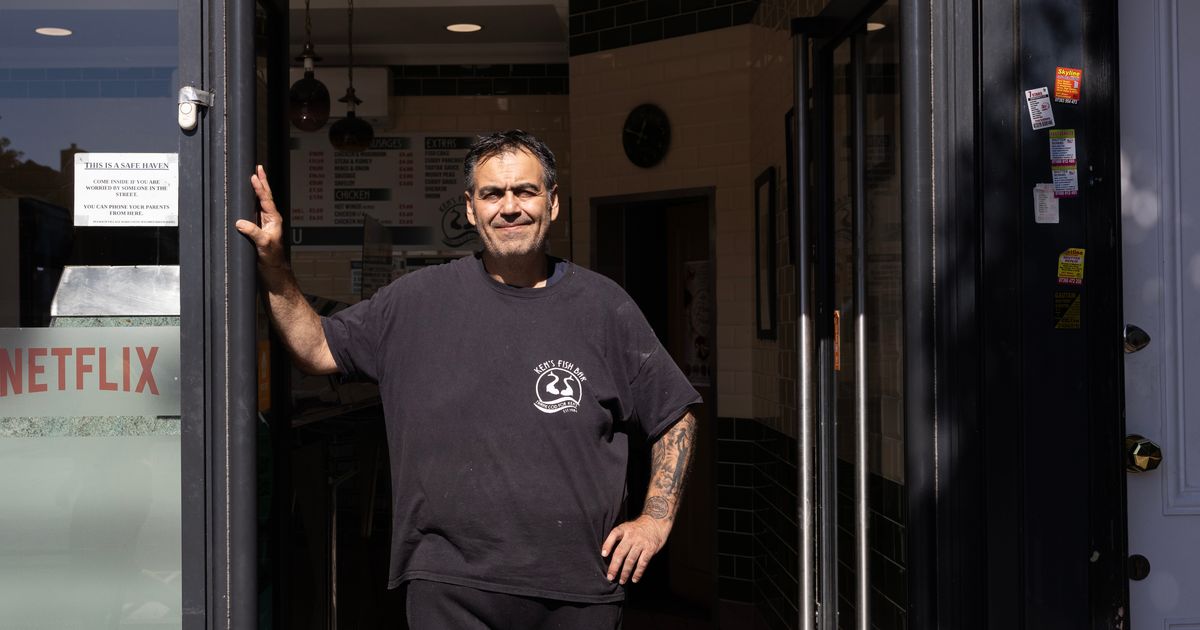 A man standing in the open doorway of a chip shop in a Ken's Fish Bar T-shirt, with a printed notice in the window beside him