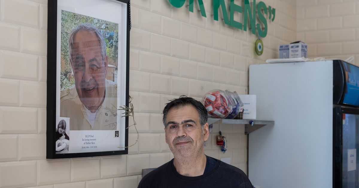 A man in a black T-shirt standing in a tiled chip shop beside a large framed photograph of an older smiling man, captioned in loving memory of Father Ken