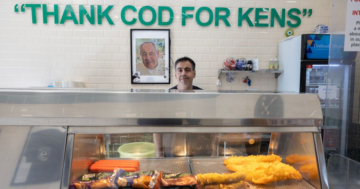 A man standing behind a fish and chip shop counter under large green letters reading Thank Cod For Kens, with his father's framed photograph on the tiled wall behind him